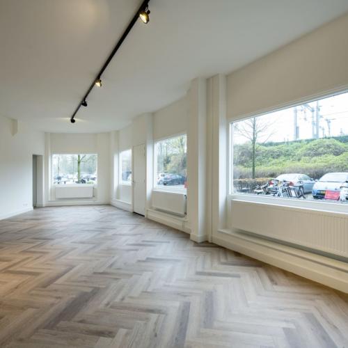 Een lichte, moderne leegstaande ruimte met chevron parket en een grote raammuur met uitzicht op groen.