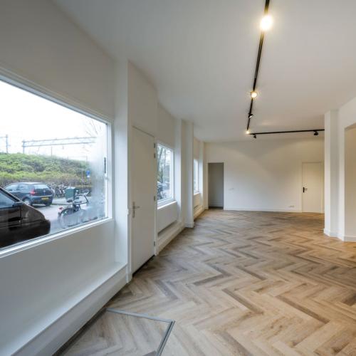 Lichte, lege hal met houten vloer, grote ramen en een moderne, minimalistische lichtarmatuur.