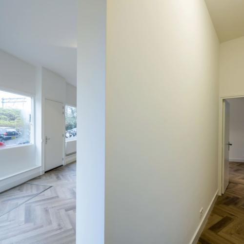 Interieurfoto van een woonkamer met groot raam en een gang die naar een andere kamer leidt.