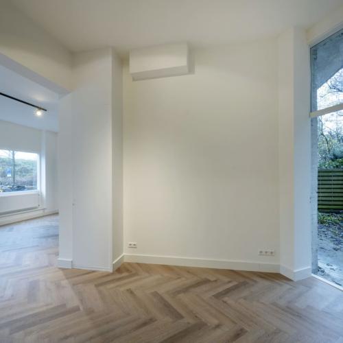 Lichte, moderne leeg kamer met houten vloer in chevronpatroon, grote ramen en een glazen deur die veel natuurlijk licht binnenlaat.