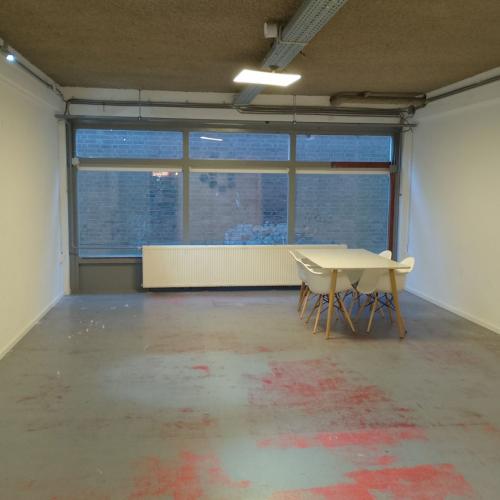 Grote, lege studio met een raamwand en een tafel, op een betonnen vloer met rood-grijs vlekken.
