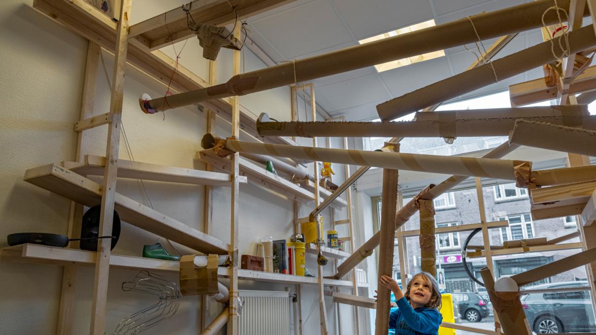 Een kind speelt met de reuzenknikkerbaan die de hele ruimte vult. De bal rolt door tapijtkokers.
