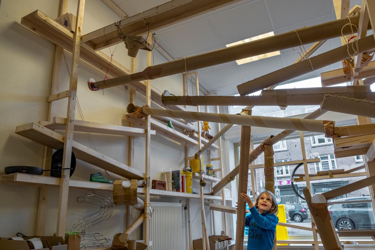 Een kind speelt met de reuzenknikkerbaan die de hele ruimte vult. De bal rolt door tapijtkokers.