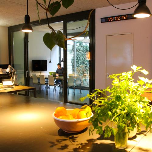 Op een donkere tafel staan een bloeiende plant en een oranje kom, met grote ramen die een modern interieur zichtbaar maken.