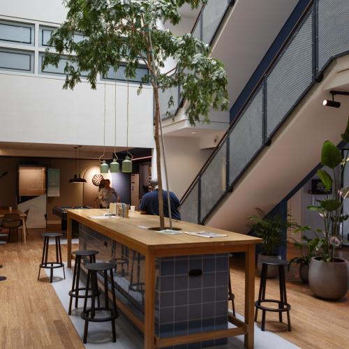 Open woon-werkruimte met een lange houten tafel en krukken. Een grote binnenboom en moderne architectuur zijn te zien.