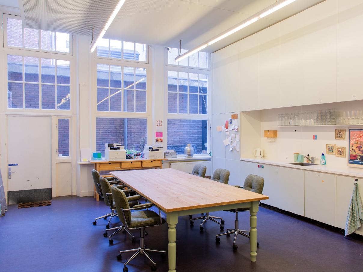 Taalhuis keuken: een grote tafel et stoelen rondom. Lang aanrecht, afwasmachine en glazen op plank. 