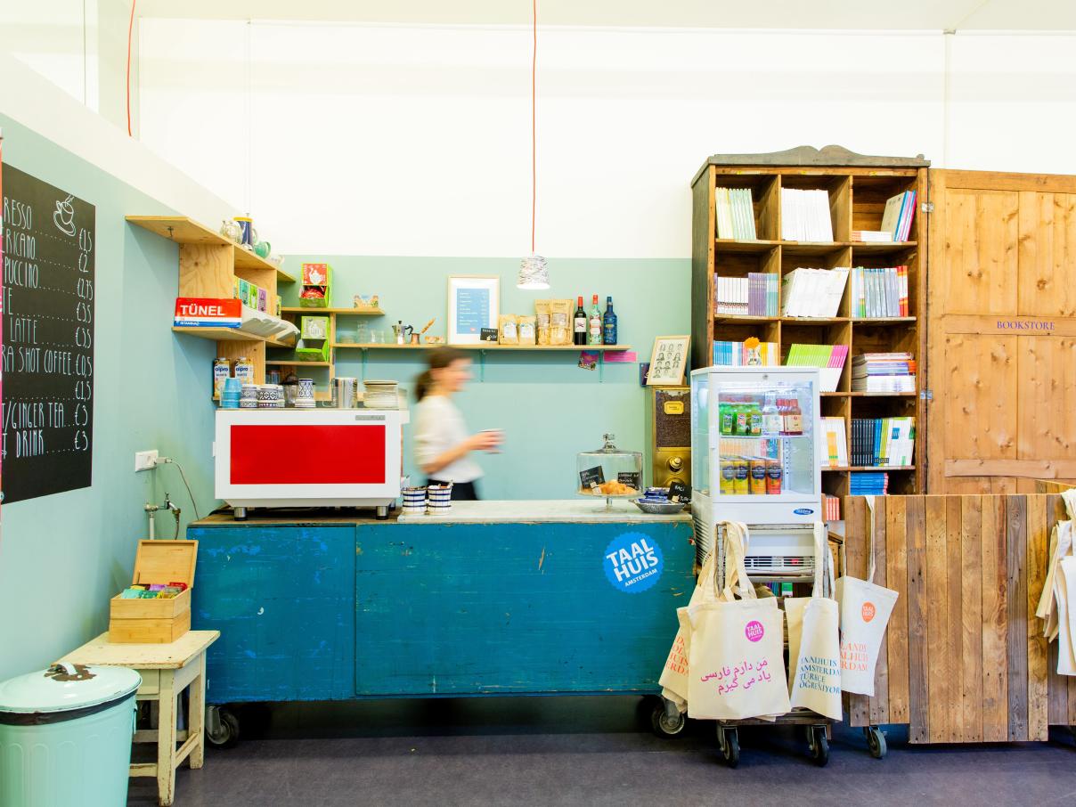 Taalhuis café: bar met koffiemachine, koelkastje met fris, vrouw achter de bar, boekenkast en tasjes
