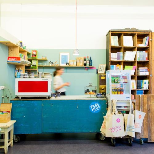 Taalhuis café: bar met koffiemachine, koelkastje met fris, vrouw achter de bar, boekenkast en tasjes