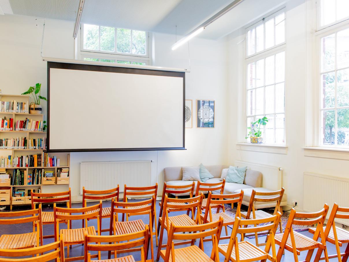 Taalhuis café: houten stoelen in bioscoopopstelling, beamer, grote ramen, boekenkastje van de bieb