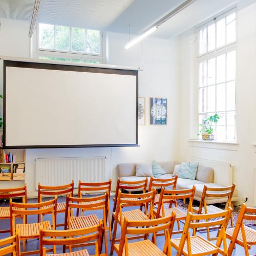 Taalhuis café: houten stoelen in bioscoopopstelling, beamer, grote ramen, boekenkastje van de bieb