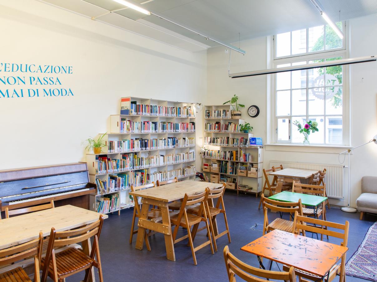 Taalhuis café: piano, 5 tafeltjes met stoelen, boekenkasten van de bieb, groot raam, bank en tapijt 