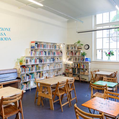 Taalhuis café: piano, 5 tafeltjes met stoelen, boekenkasten van de bieb, groot raam, bank en tapijt 
