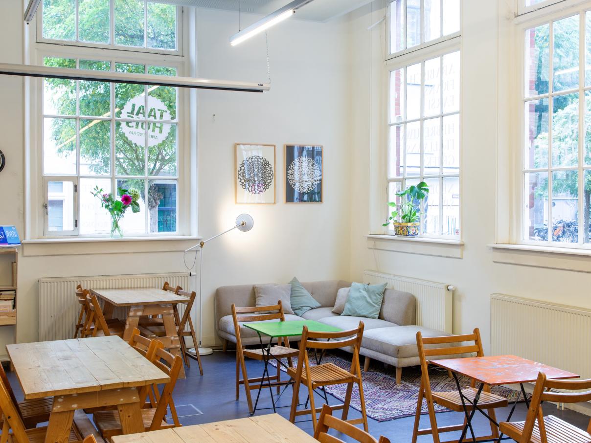 Taalhuis café: tafeltjes met stoelen, drie grote ramen, een hoekbank, schilderijen, klok, plantjes