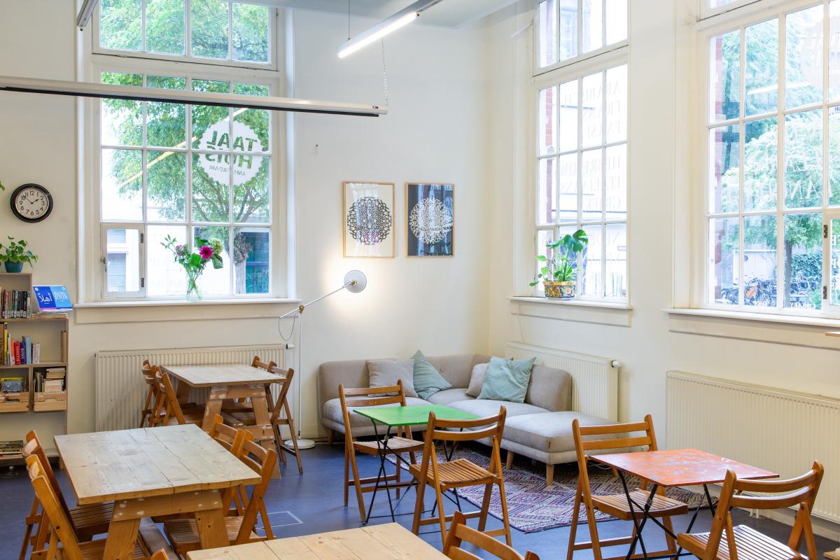 Taalhuis café: tafeltjes met stoelen, drie grote ramen, een hoekbank, schilderijen, klok, plantjes