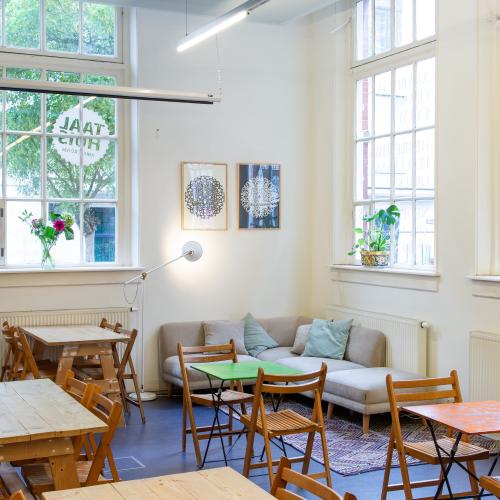 Taalhuis café: tafeltjes met stoelen, drie grote ramen, een hoekbank, schilderijen, klok, plantjes
