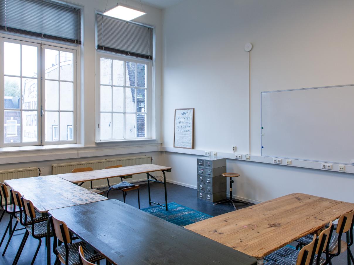 Taalhuis grote ruimte: lokaal, ramen, grote tafels in u-vorm. Stoelen, een archiefkastje, whiteboard