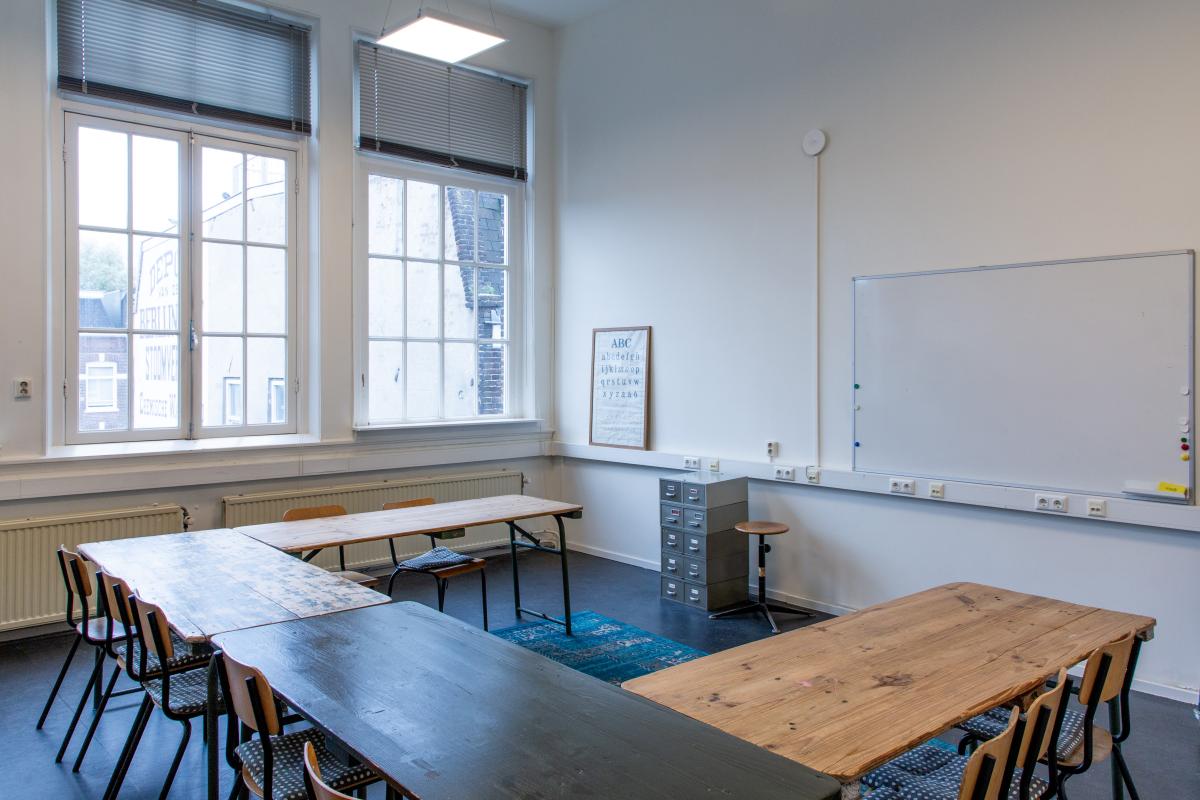 Taalhuis grote ruimte: lokaal, ramen, grote tafels in u-vorm. Stoelen, een archiefkastje, whiteboard