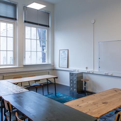 Taalhuis grote ruimte: lokaal, ramen, grote tafels in u-vorm. Stoelen, een archiefkastje, whiteboard