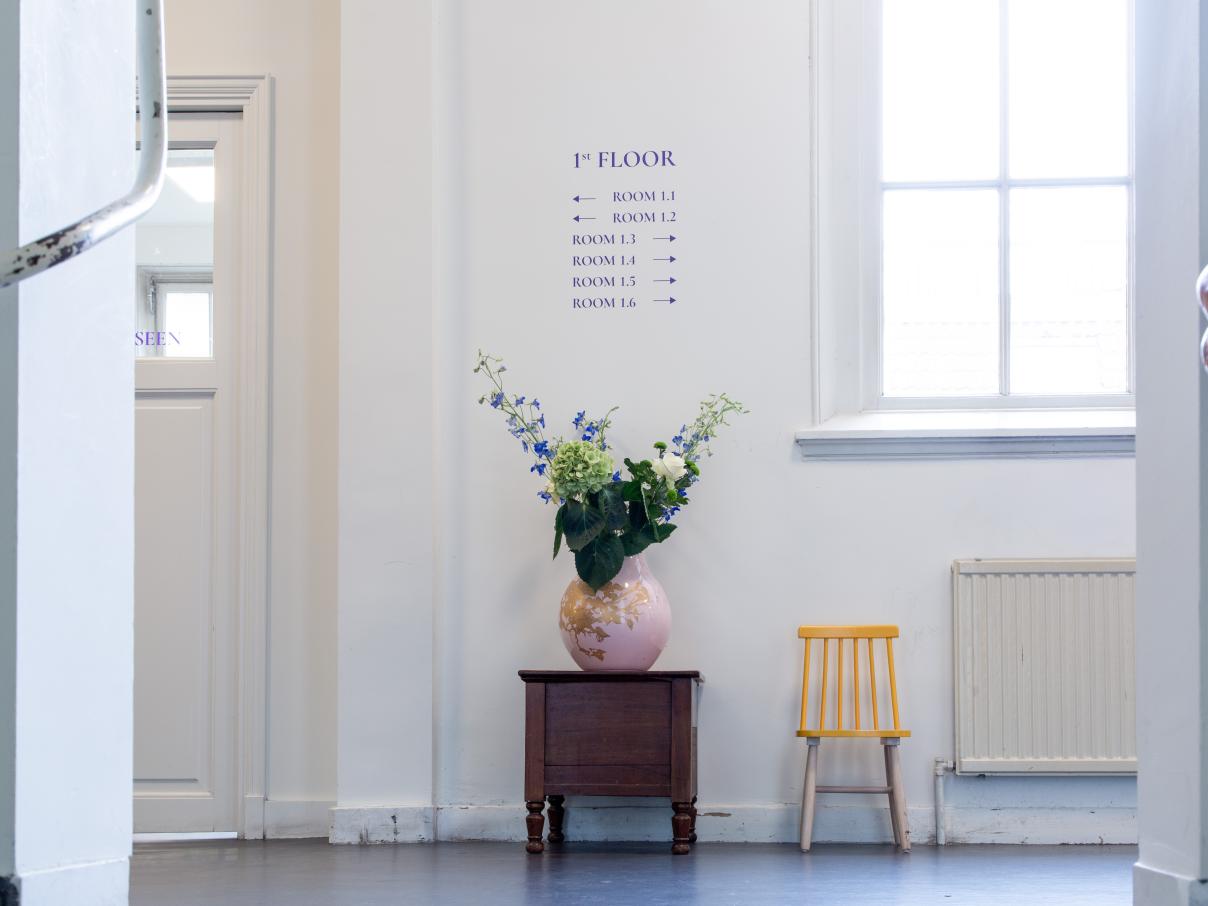 Een lichte hal, gezien vanaf de trap, bloemen in een vaas en een geel stoeltje. Bewegwijzering. 