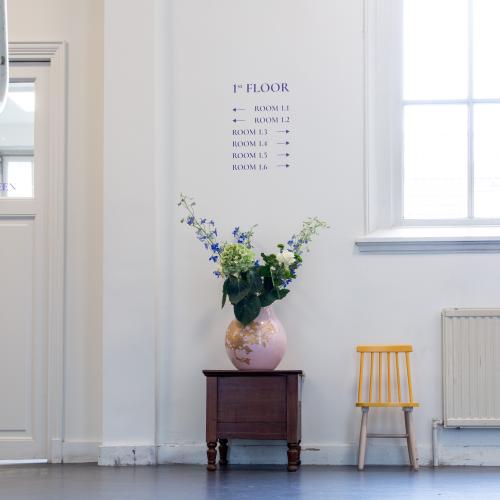Een lichte hal, gezien vanaf de trap, bloemen in een vaas en een geel stoeltje. Bewegwijzering. 