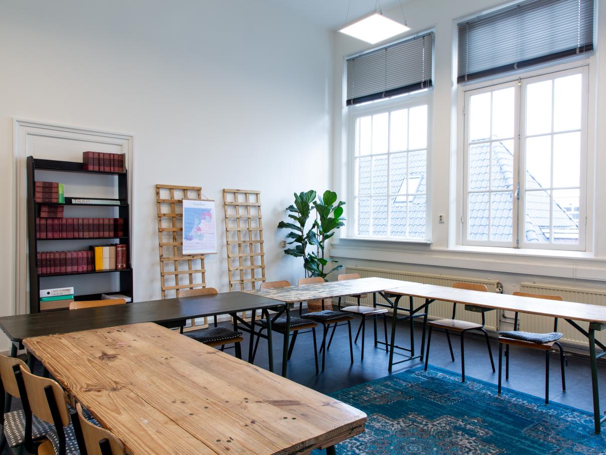 Licht lokaal met grote tafels in u-vorm, boekenkast en plant op de achtergrond. 