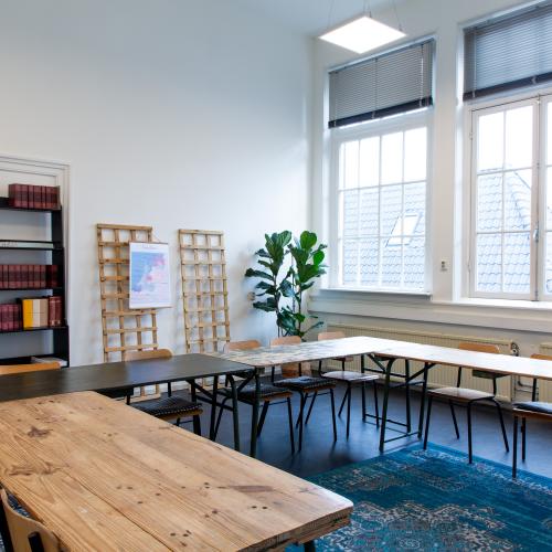 Licht lokaal met grote tafels in u-vorm, boekenkast en plant op de achtergrond. 