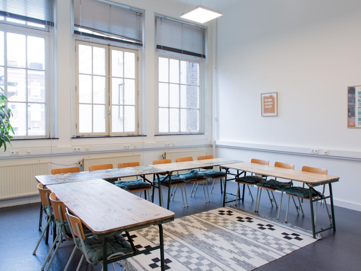 Groot licht lokaal, stoelen en grote tafels in u-vorm. Posters aan de muur. Plant in de hoek. 