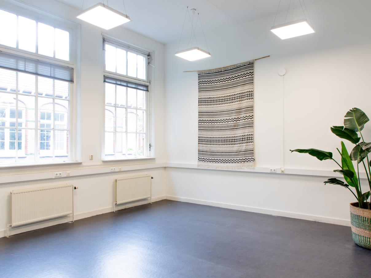 Grote ruimte zonder tafels/stoelen. Plant in de hoek en een wandkleed aan de muur.