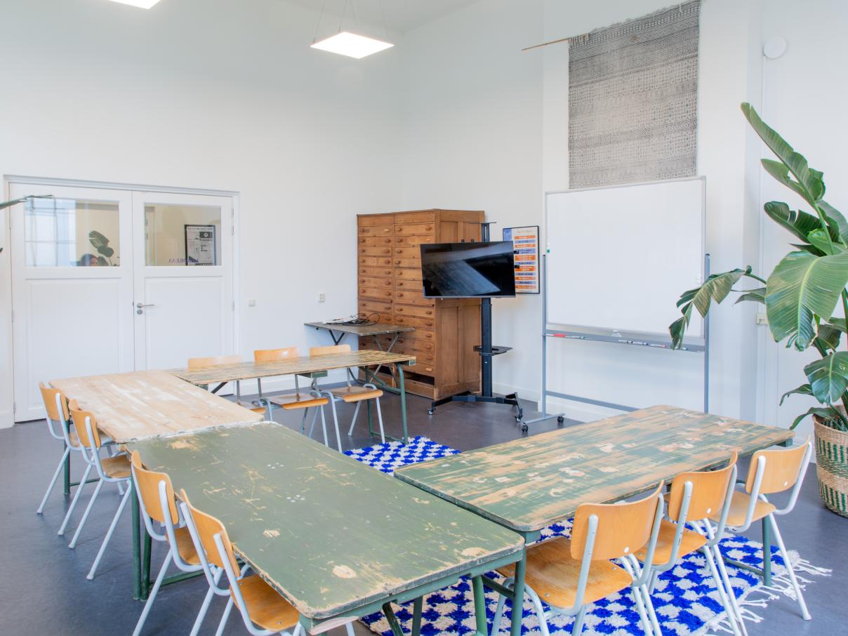 Grote ruimte met tafels en stoelen in u-vorm. Planten in de hoek. Whiteboard en een groot tv-scherm