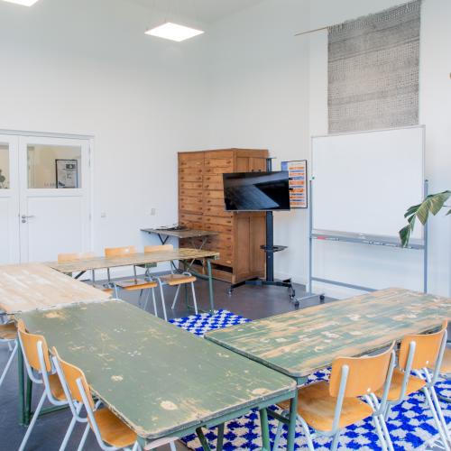 Grote ruimte met tafels en stoelen in u-vorm. Planten in de hoek. Whiteboard en een groot tv-scherm