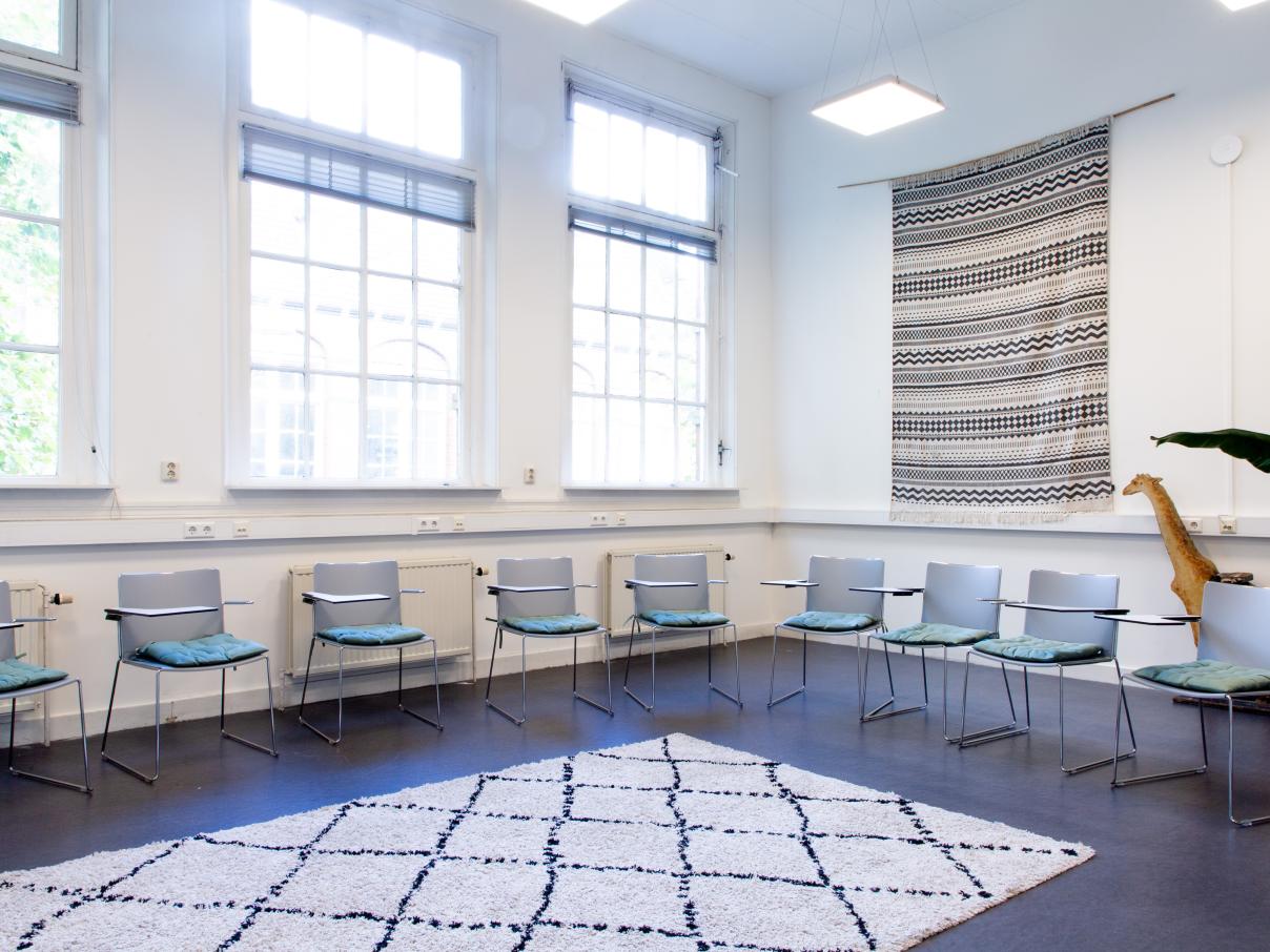 Grote ruimte met stoelen in een kring. Stoelen hebben kleine klaptafeltjes. Zacht kleed op de vloer.