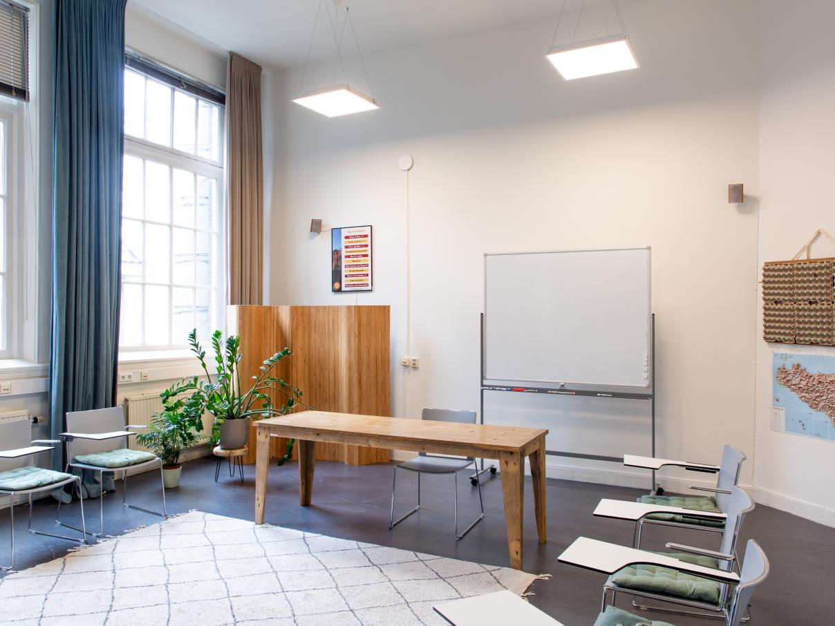 Ruimte met hoog plafond, stoelen in een kring. Een bureau voor een whiteboard. Posters aan de muur.