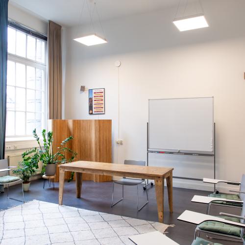 Ruimte met hoog plafond, stoelen in een kring. Een bureau voor een whiteboard. Posters aan de muur.