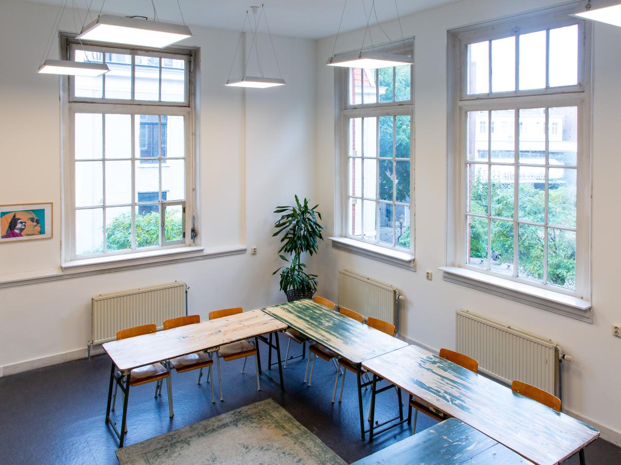 Hoge ruimte met tafels in u-vorm. Drie grote ramen en een plant in de hoek.