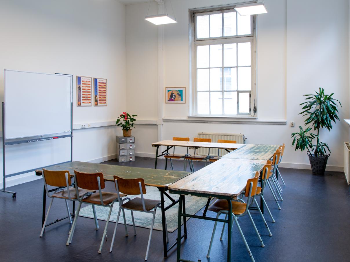 Grote ruimte met een whiteboard, grote tafels en stoelen, archiefkastje en planten in de hoek.