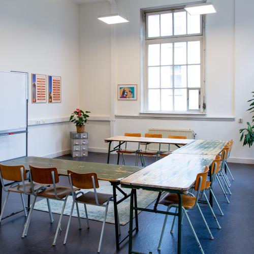Grote ruimte met een whiteboard, grote tafels en stoelen, archiefkastje en planten in de hoek.