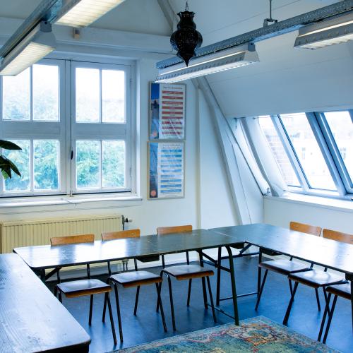 Een lokaal met rijen tafels en stoelen, badend in natuurlijk licht van grote raampartijen.
