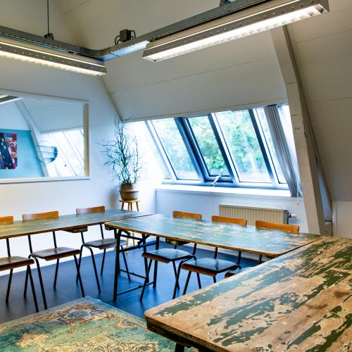 Diverse lange, ruwe tafels en stoelen in een lichte, moderne vergaderruimte met grote ramen.