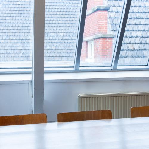 Grote raampartij met een zicht op bakstenen gebouwen, met een bank en tafel op de voorgrond.