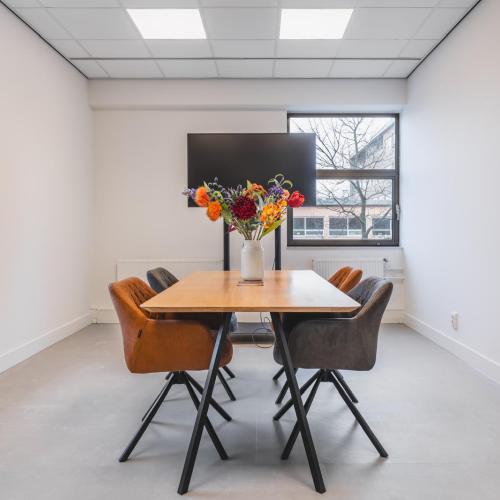 Modern vergaderruimte met een houten tafel, vier stoelen en een vaas met bloemen.