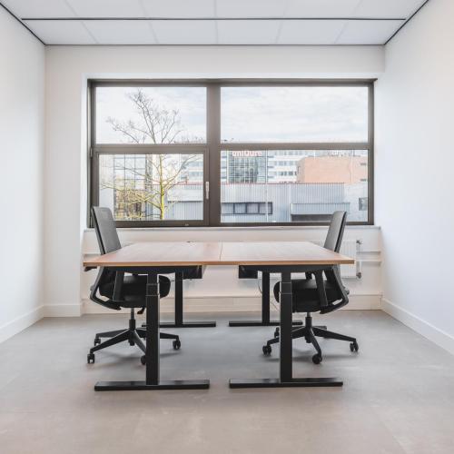Modern kantoor met twee bureaus en stoelen voor twee personen, geplaatst onder een groot raam.