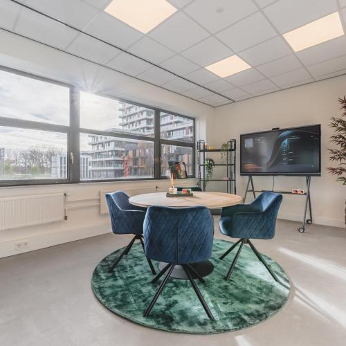 Moderne vergaderruimte met ronde tafel en donkerblauwe stoelen op een groen fluwelen kleed. Het raam en de tv zijn zichtbaar.