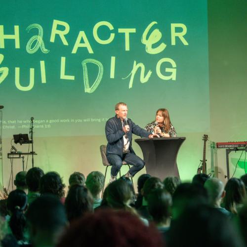 Man en vrouw op podium praten voor een groot, groen bord met de tekst "CHARACTER BUILDING".