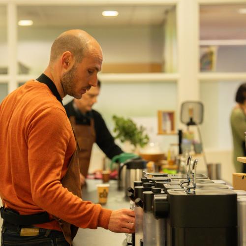 Een man in oranje trui serveert drankjes bij een buffet. Achter hem staan gasten in een lichte zaal.