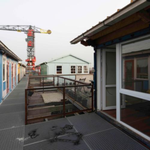 Een loopbrug tussen gekleurde gebouwen en ramen, met op de achtergrond een grote bouwkraan die de lucht domineert.