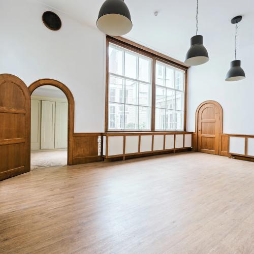 Een grote, lege zaal met lichte houten vloer, witte muren, grote ramen en klassieke houten deuren.