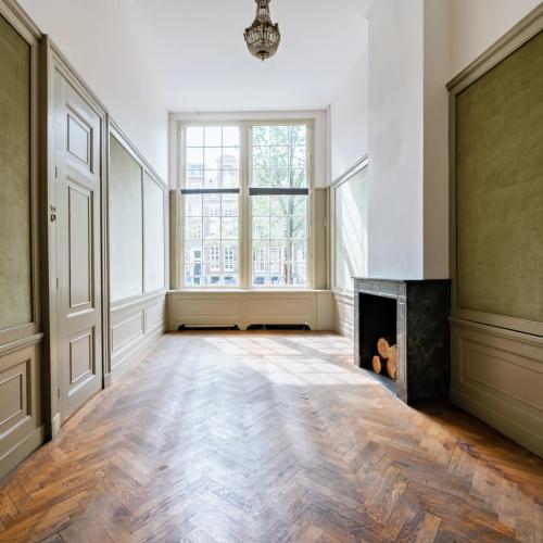 Grote woonkamer met houten vloer, hoog raam en wandpanelen in salgroen.