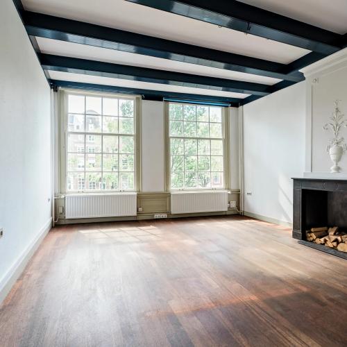 Licht binnen via grote ramen; de kamer heeft een houten vloer en een donker plafond.