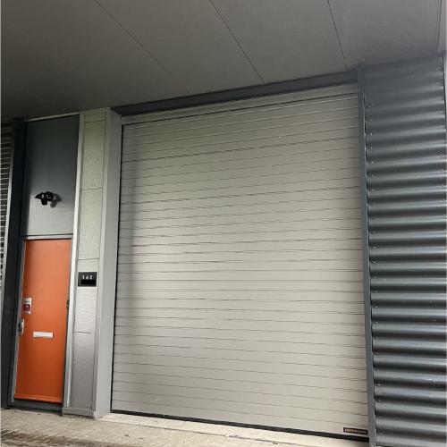 Gesloten lichtgrijze, geribbelde garagedeur in een commerciële ingang. Links staat een oranje paneel en rechts is een metalen poort te zien.