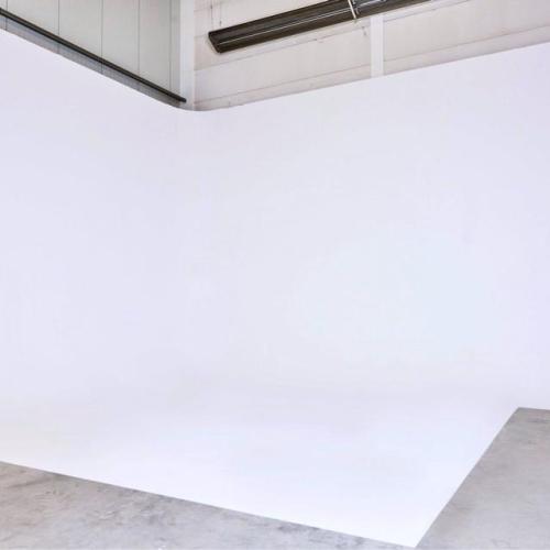 Witte, lege hoek van een binnenruimte met een betonnen vloer en lichte, plintenachtige architectuur.